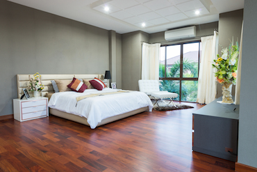 a large room with a hardwood floor, a tall window overlooking trees, a two-person bed with a single beside table, and a padded chair next to the window